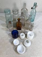 Front view photo showing an assortment of vintage antique glass bottles and jars in different colors, including clear, amber, aqua green, and cobalt blue with wire bail closure on one bottle and various white milk glass jars in front.