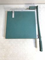 Top-down view of vintage photo cutter showing green grid cutting surface, white ruler guide, and long metal lever arm with green handle and mounted blade assembly.