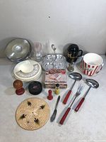 Vintage kitchen items displayed on table including ladles, condiment holders, measuring cup, grater, wooden egg cup, woven trivet, and popcorn bucket.