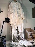 Full view of hanging white military snow camouflage coat and pants set with boots standing below. A pair of white glove and rucksack covers are placed around the boots.