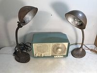 Two vintage metal gooseneck table lamps and one vintage Philips tabletop radio displayed on a flat surface. Lamps show rust and visually damaged electrical cord on one lamp, radio has weathered blue-green metal casing.