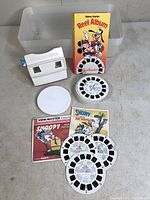 View of entire lot showing View-Master viewer, white round slide case, Disney Stories Reel Album, Snoopy slide booklets and reels, and a plastic bin for storage.
