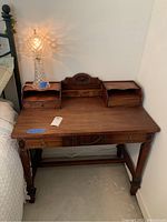 Full view of antique wooden desk with center drawer, two cubby drawers on top, and crystal glass lamp placed on the right side.