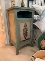 Front angled view showing green painted cabinet with topiary painting on the door, open shelf above, and decorative side panels.