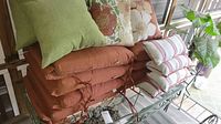 Shows multiple chair cushions stacked on a metal rack on a porch. Includes various colors and patterns such as rust-colored, striped red and white, floral, and green cushions.