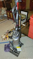 Full view of the Dyson DC25 vacuum cleaner upright on carpet showing body, hose, and handle in gray, silver, and red with some accessories at base.