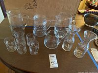 Photo showing four pitchers including one etched with floral design, five matching etched glasses, and two clear glass carafes on a table. The tallest pitcher measures about 8.5 inches.