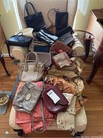 Overview of 16 assorted handbags in leather, faux leather and canvas, stacked on an armchair and ottoman showing the scale, colors, and styles.