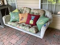 Full view of wicker hanging swing with floral cushions and colorful decorative pillows on outdoor brick patio.