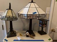 Three table lamps on a white surface, showing different stained glass style shades and lamp base styles. One shade appears cracked.