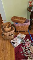 Set of woven Longaberger baskets and catalogs on wooden floor near a floral rug and a round wooden table.