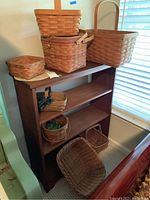 Eight woven baskets displayed on and beside a wooden bookshelf in natural light. Various sizes and shapes clearly visible, including baskets with handles and lids.