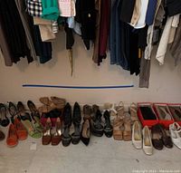 Wide shot showing three rows of assorted women's shoes including flats, sandals, and heels in neutral colors and bright green and orange, arranged on floor below hanging clothes.