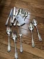 Overhead on wood floor showing mixed flatware pieces in three patterns, including monogrammed sterling and silver plate spoons, forks, knives.