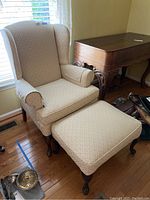 View of the wingback chair and ottoman together near a window with wood flooring and a wooden furniture piece nearby
