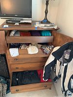 Wooden dresser with three drawers open showing an assortment of winter hats, headbands, gloves, sweatshirts, snow/ski pants, and change purses stacked inside. TV and lamp on top of dresser.