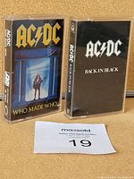 Two AC/DC audio cassette tapes in plastic cases standing upright, titles visible: 'Who Made Who' and 'Back in Black'.