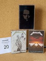 Front view of two Metallica cassettes, '...And Justice for All' and 'Master of Puppets', plus a third cassette with black cover showing a face.