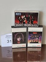 Three KISS audio cassette tapes placed on a reflective surface. Visible titles: 'Smashes, Thrashes & Hits', 'Love Gun', and 'The Priceless Collection'.