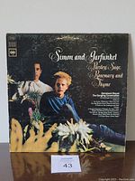 Front cover of Simon and Garfunkel vinyl album Parsley Sage Rosemary and Thyme showing both artists seated outdoors with foreground flowers.