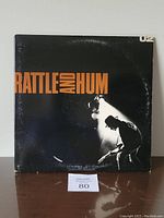 Front cover of U2 Rattle and Hum vinyl LP showing title in orange and white text, black background and silhouette of guitarist.