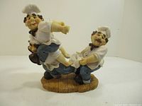 Side angle view of ceramic bottle holder showing two chefs, one carrying the other holding a bottle, on a decorative base.