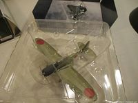 Close-up image of die cast model plane inside clear plastic packaging. Shows details of olive green paint, red roundels on wings, propeller fine, and cockpit canopy.
