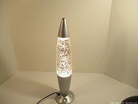 Silver glitter lava lamp with conical top, round base, and electrical cord, photographed with bright lighting to show details.