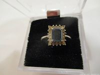 Close-up front view of the ring showing central blue gemstone and surrounding clear stones in display box.