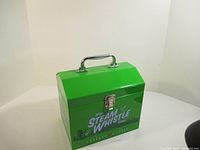Photo of a green metal Steam Whistle lunch box with silver handle and clasp, showing front branding, against white background.