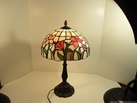 Full view of the lamp illuminated showing the stained glass shade with floral pattern and the dark ornate metal base