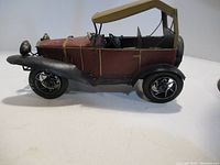 Side view of the vintage style ornamental tin car showing maroon body with black fenders and wheels, and tan convertible roof.