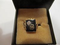 Front view of the vintage gold tone ring with rectangular black onyx stone, diamond-shaped clear stone, and gold letter 'S'. Ring displayed on beige cushion inside a black ring box.