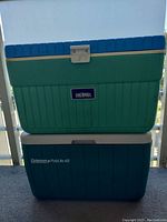 Two coolers stacked: top is a green Thermos cooler with blue lid and silver latch, bottom is a blue Coleman PolyLite 48 cooler.