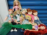 Photo showing nine assorted dolls including Ariel mermaid dolls, plush dolls and porcelain style doll arranged against blue background.