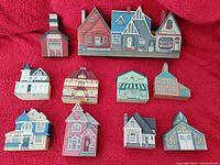 Photo showing 12 hand painted miniature wooden buildings arranged in a grid on red fabric background.