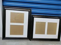 Photo showing two large wooden picture frames with matting inside, black-finished frames, seated on the floor.