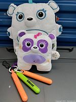 Photo showing two Inkoos washable toy drawing pads, one large white bear-like and one small purple and white dog-like, with two colorful washable pens (orange and green) in front.