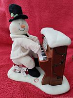 Snowman playing a piano with Christmas lights decoration, showing the item on a red fabric background.
