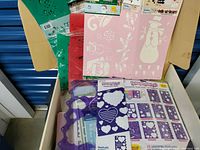 Multiple seasonal and holiday-themed stencils in red, green, pink, purple, and other colors in a cardboard box.
