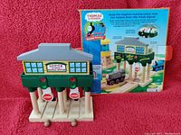 Front view of the wooden Sodor Signal House with gray roof, green and red detailing, and hanging stop signs. Blue packaging backing behind showing product description and assembly instructions.