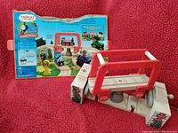 Photo showing Thomas & Friends rotating bridge playset with two ascending tracks in front of original packaging box, all items placed on a red textured cloth surface.