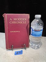 Front cover of the book 'A Modern Chronicle' by Winston Churchill showing red cloth cover with gold lettering, placed on a book stand next to a water bottle.