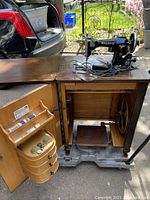 Overall view of the Singer Phoenix sewing machine mounted on the original wooden cabinet with open door revealing storage compartments and foot pedal.
