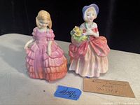 Photo of two vintage Royal Doulton figurines modeled as young women with detailed dresses, one holding a flower basket, both in pastel colors.