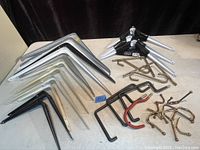 A variety of metal wall shelf brackets in black, white, and chrome finishes arranged on a flat surface, alongside assorted metal hooks including brass and chrome types with some hooks having red rubber grips.