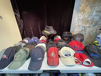 First photo showing baseball caps and one Australian wool hat on a blue table against a dark backdrop and stone wall.