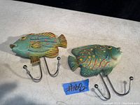 Photo of the pair of fish-shaped towel holders with painted colorful details and metal dual hooks against a light background.