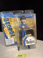 New in box George Harrison figurine from The Beatles by McFarlane Toys with blue outfit and guitar, packaging shows The Beatles logo.