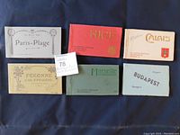 Six antique postcard booklets arranged on a surface showing front covers and a label with lot information